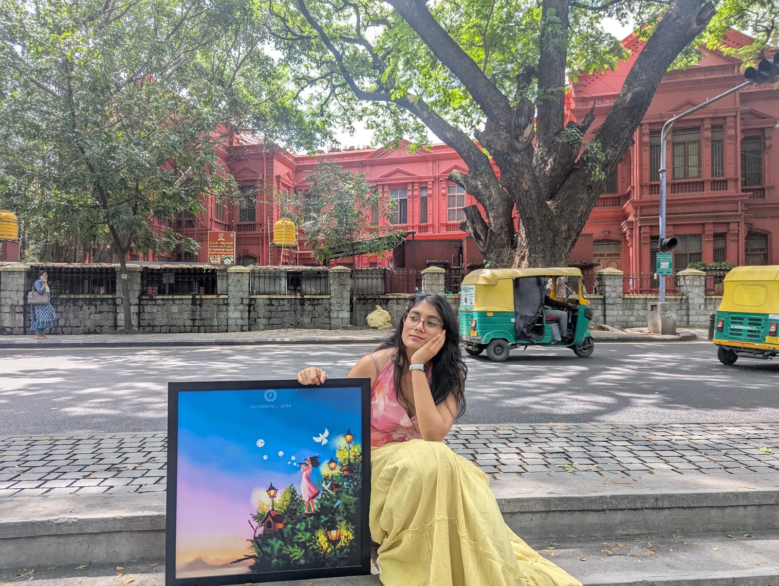 Saudamini Jena seated beside a framed painting with a red building, trees, and auto rickshaws in the background.